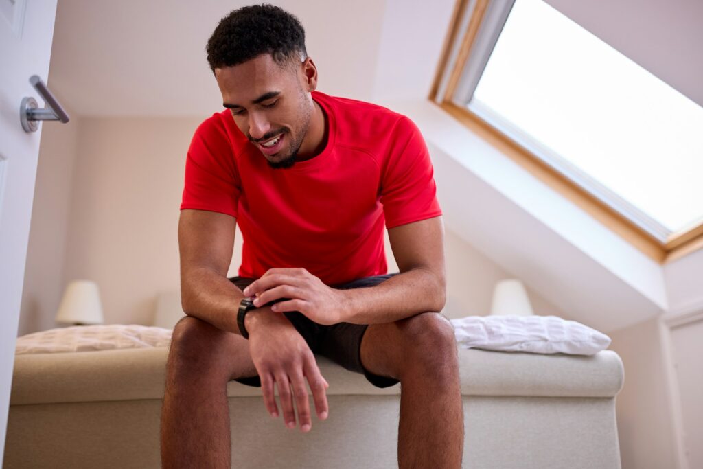 Young Man In Bedroom At Home In Fitness Clothing Checking Fitness App On Smart Watch Before Exercise