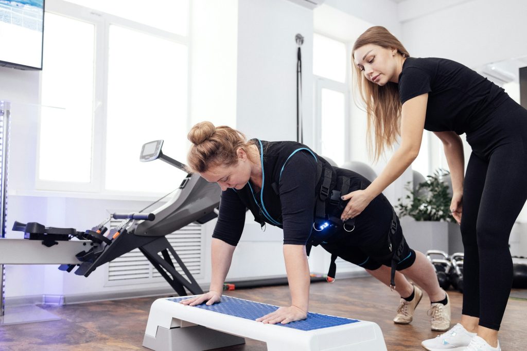 Plump woman middle age in ems suit is engaged on step platform with personal fitness trainer.